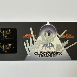 May include: A black card featuring two film stills from Stanley Kubrick's "Clockwork Orange." The card has a white triangle with an eye graphic and the film's title. The card is signed in silver ink.