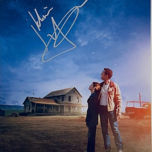 May include: A movie poster featuring a night sky with a signature. Two people stand in front of a house, looking up. The sky is a deep blue with stars and clouds. The house is old and weathered.