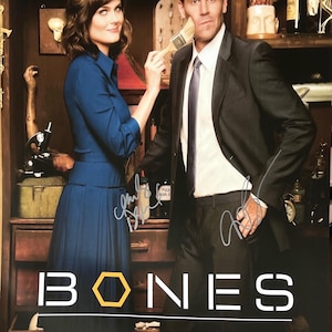 May include: A signed poster of the TV show "Bones" featuring the main characters, a woman in a blue dress and a man in a suit, standing in front of a shelf with various items. The poster has the show's title in large white letters with a yellow hexagon.