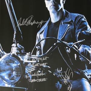 May include: Movie poster for Terminator 2: Judgment Day featuring Arnold Schwarzenegger on a motorcycle, holding a shotgun. The poster has signatures and the text "It's Nothing Personal."