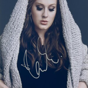 Puede incluir: Retrato de una persona con un suéter con capucha gris sobre una camiseta negra. La persona tiene el pelo largo, ondulado y castaño y está mirando hacia abajo. El suéter es de punto grueso. La camiseta tiene una firma blanca.