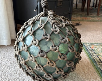 May include: A large, round, green glass buoy with a thick, brown rope net. The buoy is hanging from the rope net.