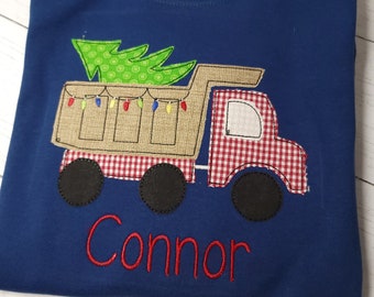 May include: Navy blue long-sleeve shirt featuring an appliqued dump truck carrying a Christmas tree. The truck bed is tan, the cab is red and white checkered, and the wheels are black. The name "Connor" is embroidered in red.