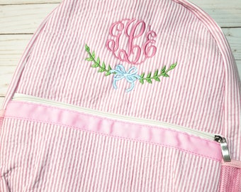 May include: A pink and white seersucker backpack featuring a front zippered pocket with a pink ribbon trim. The upper section displays an embroidered pink monogram with a light blue bow and green leafy vine accents. A pink mesh side pocket is visible.