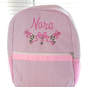 Pode incluir: Uma mochila de seersucker rosa e branca com o nome Nora bordado em letra cursiva rosa. Abaixo do nome, há um desenho bordado de flores rosas com folhas verdes e um laço rosa. A mochila possui um bolso frontal com zíper e malha nas laterais.