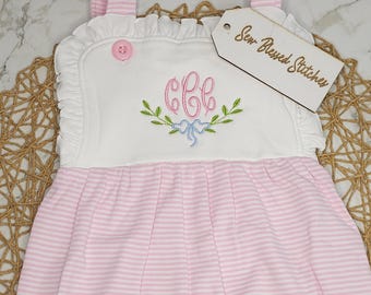 May include: A white and pink striped romper featuring a white bodice with ruffled edges, a pink button, and an embroidered monogram with green leaves and a blue bow. A wooden tag attached to the shoulder displays the text Sew Blessed Stitches.