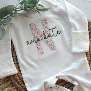 May include: White baby sleeper with long sleeves and snap closures. Features a floral-patterned letter "N" and the name "nora kate" embroidered in green thread. The sleeper is made of soft fabric.