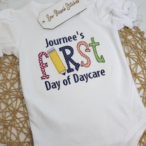 K&ouml;nnte beinhalten: Wei&szlig;er Strampler mit dem Text "Journee's First Day of Daycare" in bunten gestickten Buchstaben. Der Buchstabe "F" ist ein Bleistift.
