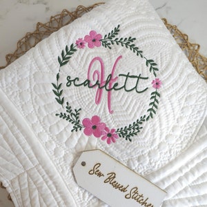 Puede incluir: Manta de bebé acolchada blanca con una corona de flores bordada con el nombre "Scarlett" y la letra "S". Una etiqueta de madera con el texto "Sew Blessed Stitches" está en la manta.