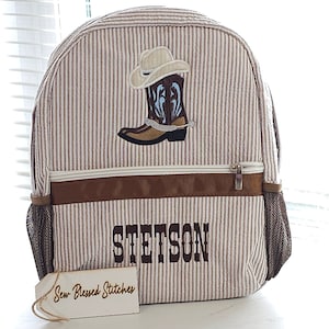 May include: A brown and white striped backpack with a brown leather trim. The backpack has a brown boot and cowboy hat embroidered on the front. The name "STETSON" is embroidered in brown on the front of the backpack. A white tag with the text "Sew Blessed Stitches" is attached to the backpack.