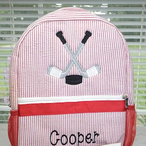 May include: Red and white striped backpack with a hockey design. It has crossed hockey sticks and a puck. The name "Cooper" is embroidered on the front. Includes a red pocket and mesh side pocket. The tag reads "Sew Blessed Stitches."