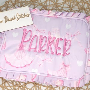 May include: A lavender baby blanket with a pink embroidered name "Parker". The blanket features a pattern of ballet shoes, bows, and hearts. The edges are trimmed with a ruffled border. A wooden sign that says "Blessed Stitches" is in the background.