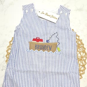 May include: A sleeveless blue and white seersucker romper featuring an embroidered brown boat with a red tackle box, a fishing rod, and a green fish. The name BRAYDEN is stitched in blue on the boat. A tag reads Sew Blessed Stitches.