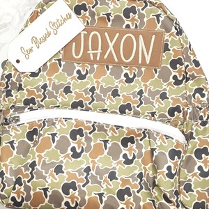 Puede incluir: Una mochila con estampado de camuflaje con el nombre "JAXON" bordado en un parche marr&oacute;n. La mochila tiene una cremallera y correas blancas, y una etiqueta que dice "Sew Blessed Stitches". El patr&oacute;n incluye tonos de marr&oacute;n, verde y negro.