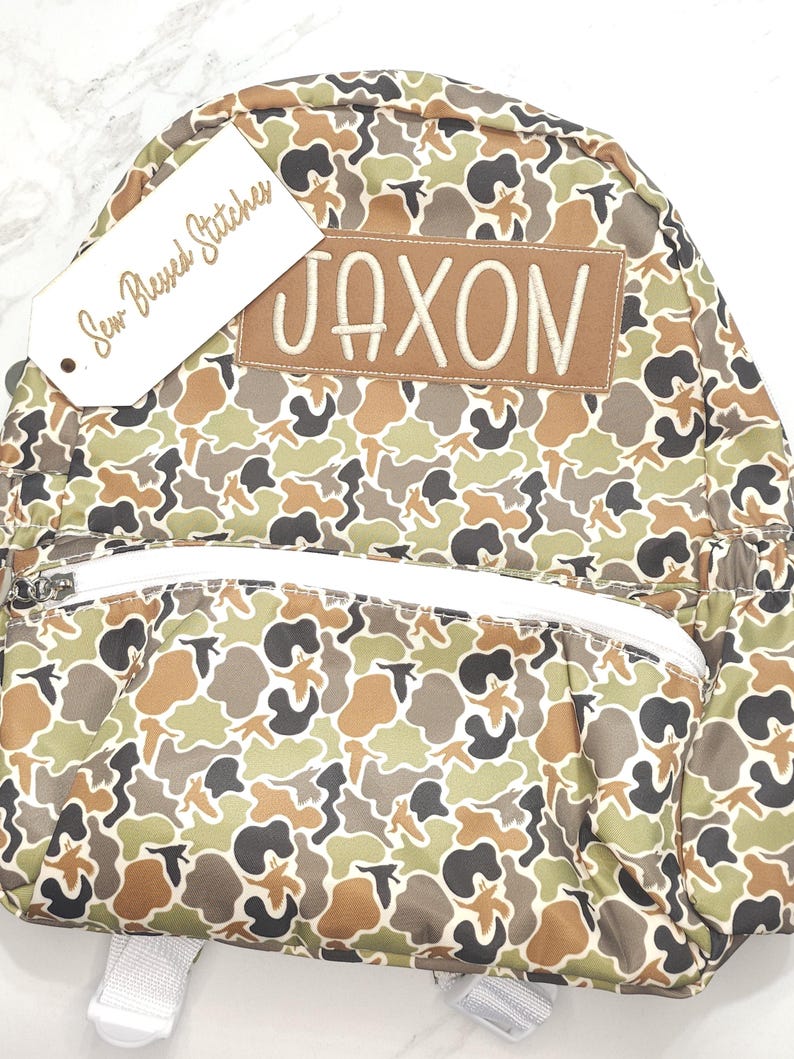 Puede incluir: Una mochila con estampado de camuflaje con el nombre "JAXON" bordado en un parche marr&oacute;n. La mochila tiene una cremallera y correas blancas, y una etiqueta que dice "Sew Blessed Stitches". El patr&oacute;n incluye tonos de marr&oacute;n, verde y negro.