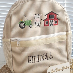Puede incluir: Una mochila de tela a rayas beige y marrón con una cremallera blanca y un bolsillo de malla. La mochila tiene imágenes bordadas de un granero rojo, un tractor verde y una vaca blanca y negra. El nombre "Emmett" está bordado en la parte delantera de la mochila. Una etiqueta de madera con el texto "Sew Blessed Stitches" está unida a la mochila.
