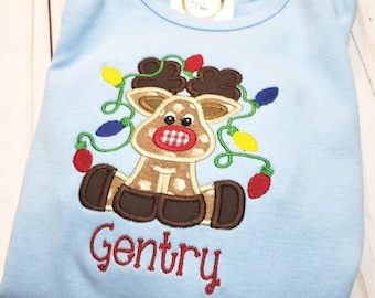 May include: A light blue baby onesie with a brown reindeer wearing a red and white checkered nose and surrounded by a string of colorful Christmas lights. The onesie is personalized with the name "Gentry" in red embroidery.