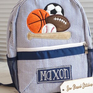 May include: Blue and white striped backpack with a navy blue trim. The backpack has a large embroidered patch with a baseball bat and various sports balls, including a baseball, basketball, football, and soccer ball. The backpack also has a smaller embroidered patch with the name "Maxon". The backpack is sitting on a white surface with a wooden tag that says "Sew Blessed Stitches".