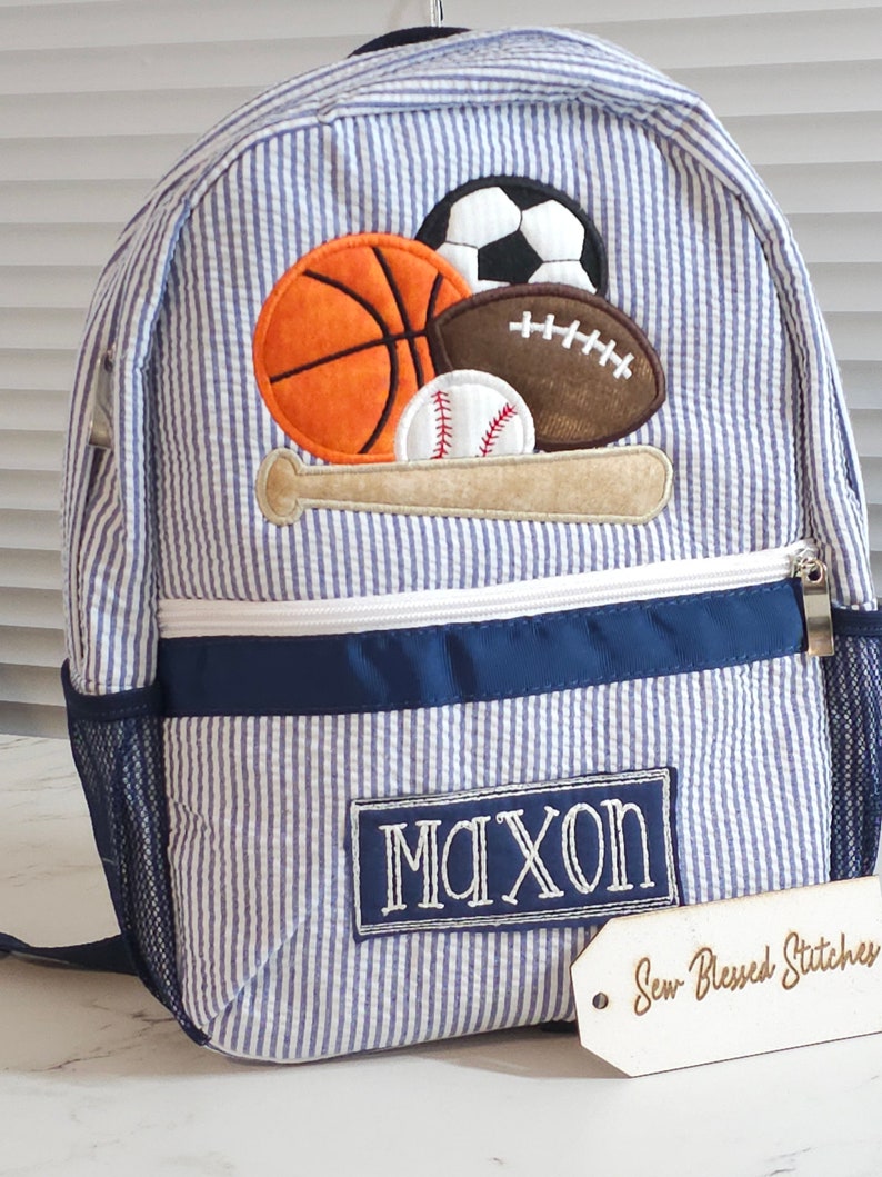 May include: Blue and white striped backpack with a navy blue trim. The backpack has a large embroidered patch with a baseball bat and various sports balls, including a baseball, basketball, football, and soccer ball. The backpack also has a smaller embroidered patch with the name "Maxon". The backpack is sitting on a white surface with a wooden tag that says "Sew Blessed Stitches".