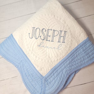 Puede incluir: Una colcha de bebé blanca y azul con un borde azul. La colcha está bordada con el nombre "JOSEPH" y la palabra "daniel" en una fuente más pequeña.
