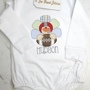 May include: White long-sleeved baby onesie with a turkey-football design. The turkey has a red helmet and holds a brown football. The name "Hudson" is embroidered below the design. A wooden tag says "Sew-Blessed Stitches".