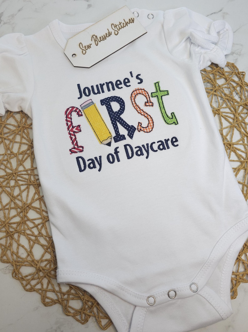 K&ouml;nnte beinhalten: Wei&szlig;er Strampler mit dem Text "Journee's First Day of Daycare" in bunten gestickten Buchstaben. Der Buchstabe "F" ist ein Bleistift.