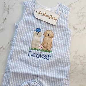 Pu&ograve; includere: Una tutina in seersucker azzurro e bianco con un ricamo raffigurante due cani, uno con un berretto blu, una palla da baseball e una mazza. Il nome Decker &egrave; ricamato in filo blu sotto i cani. Un'etichetta riporta la scritta Sew Blessed Stitches.