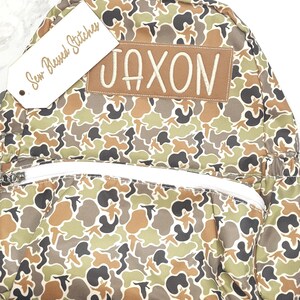 Puede incluir: Una mochila con estampado de camuflaje con un bolsillo frontal con cremallera y correas ajustables. La mochila presenta un parche marr&oacute;n con el nombre "JAXON" bordado en blanco. Una etiqueta que dice "Sew Blessed Stitches" est&aacute; adjunta.