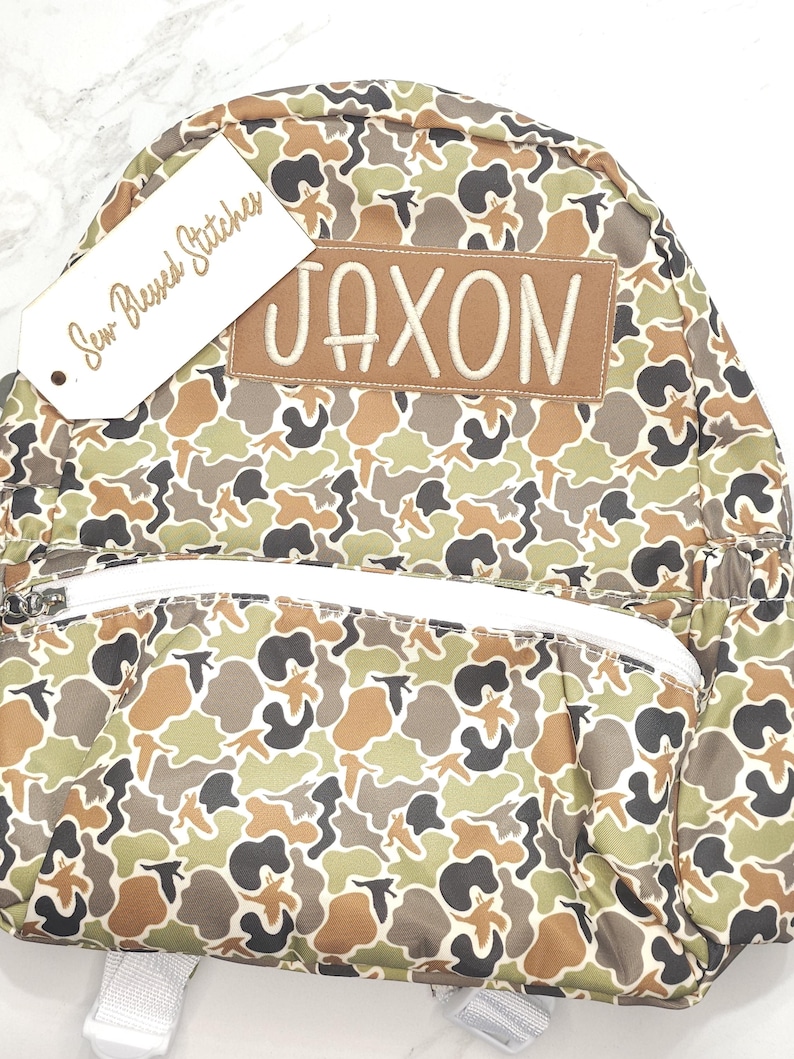 Puede incluir: Una mochila con estampado de camuflaje con un bolsillo frontal con cremallera y correas ajustables. La mochila presenta un parche marr&oacute;n con el nombre "JAXON" bordado en blanco. Una etiqueta que dice "Sew Blessed Stitches" est&aacute; adjunta.