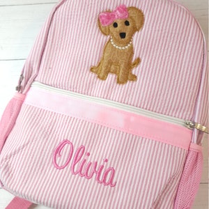 May include: Pink and white striped backpack with a brown dog embroidered on the front. The dog is wearing a pink bow and a pearl necklace. The backpack has a pink zipper and a mesh pocket on the side. The name "Olivia" is embroidered on the front of the backpack.