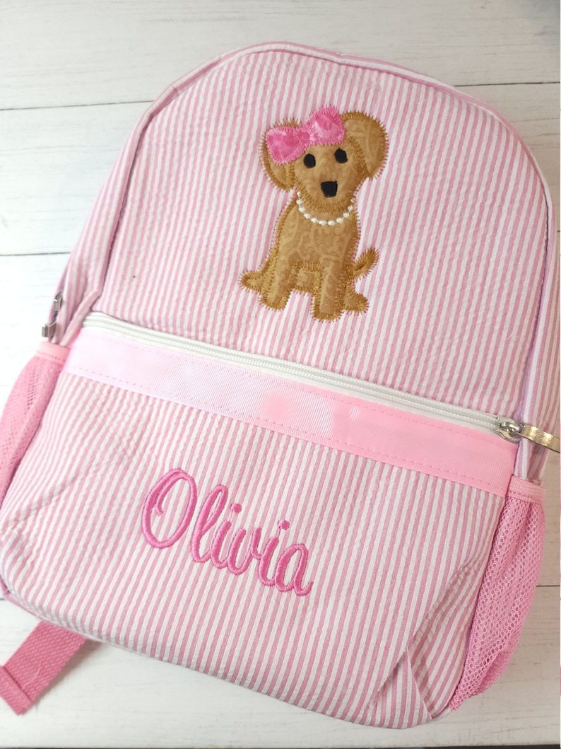 May include: Pink and white striped backpack with a brown dog embroidered on the front. The dog is wearing a pink bow and a pearl necklace. The backpack has a pink zipper and a mesh pocket on the side. The name "Olivia" is embroidered on the front of the backpack.