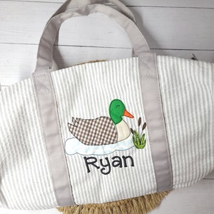 May include: A gray and white striped tote bag with a duck applique. The duck has a green head, brown and white checkered body, and an orange beak. The name "Ryan" is embroidered below the duck in black script. The bag has gray handles.