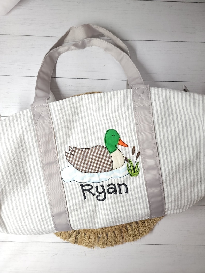 May include: A gray and white striped tote bag with a duck applique. The duck has a green head, brown and white checkered body, and an orange beak. The name "Ryan" is embroidered below the duck in black script. The bag has gray handles.