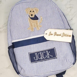 May include: Blue and white striped backpack with a dog applique and the name "JACK" embroidered on a navy blue patch. The backpack has a mesh side pocket and a tag that reads "Sew-Blessed Stitches".