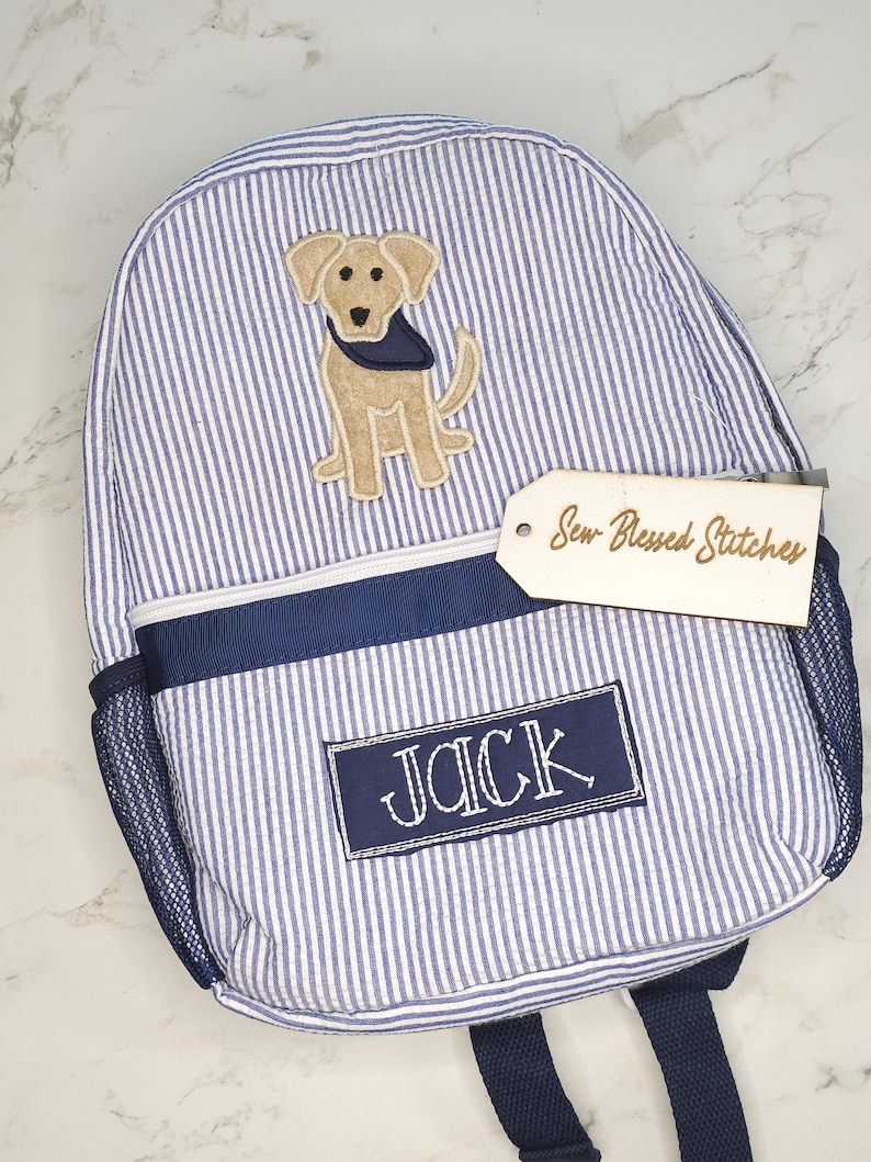 May include: Blue and white striped backpack with a dog applique and the name "JACK" embroidered on a navy blue patch. The backpack has a mesh side pocket and a tag that reads "Sew-Blessed Stitches".