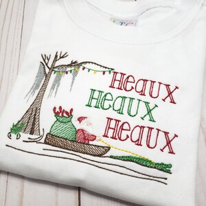 May include: A white t-shirt featuring an embroidered design of Santa Claus in a brown canoe pulling a green alligator with a yellow rope. The scene includes a cypress tree with Spanish moss and Christmas lights, with the text Heaux Heaux Heaux.