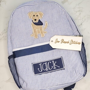May include: Blue and white striped backpack with a navy blue trim and mesh side pocket. Features an embroidered patch of a tan dog wearing a navy blue bandana. The name "JACK" is embroidered on a navy blue patch. A wooden tag reads "Sew-Blessed Stitches."