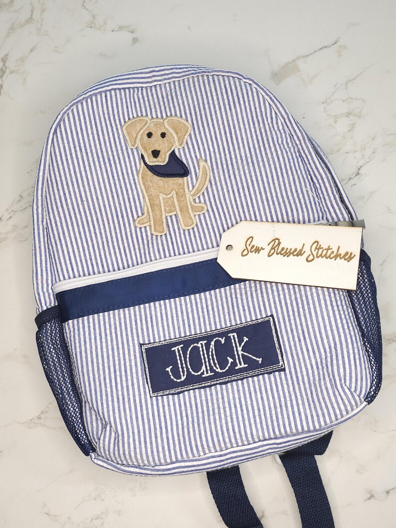 May include: Blue and white striped backpack with a navy blue trim and mesh side pocket. Features an embroidered patch of a tan dog wearing a navy blue bandana. The name "JACK" is embroidered on a navy blue patch. A wooden tag reads "Sew-Blessed Stitches."