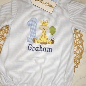 May include: A light blue and white striped baby romper with a giraffe and the number 1 embroidered on the front. The giraffe is yellow with brown spots and holds a green balloon. The name "Graham" is embroidered below.