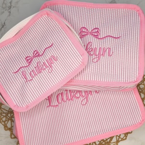 May include: Three pink and white striped cosmetic bags with the name "Laikyn" embroidered in cursive script. Each bag has a pink border and a bow design. The bags are arranged on a woven mat.
