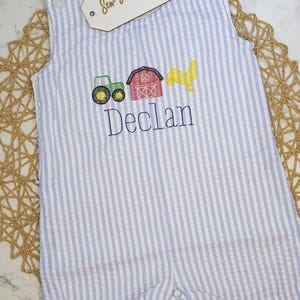 May include: Blue and white striped seersucker romper with embroidered farm scene and the name "Declan". The romper features a green tractor, red barn, and yellow rooster. The tag reads "Sew Blessed Stitches".