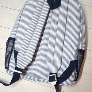 May include: A blue and white striped backpack with navy blue straps and mesh side pockets. The backpack has adjustable straps and white plastic buckles. The fabric has a vertical stripe pattern.