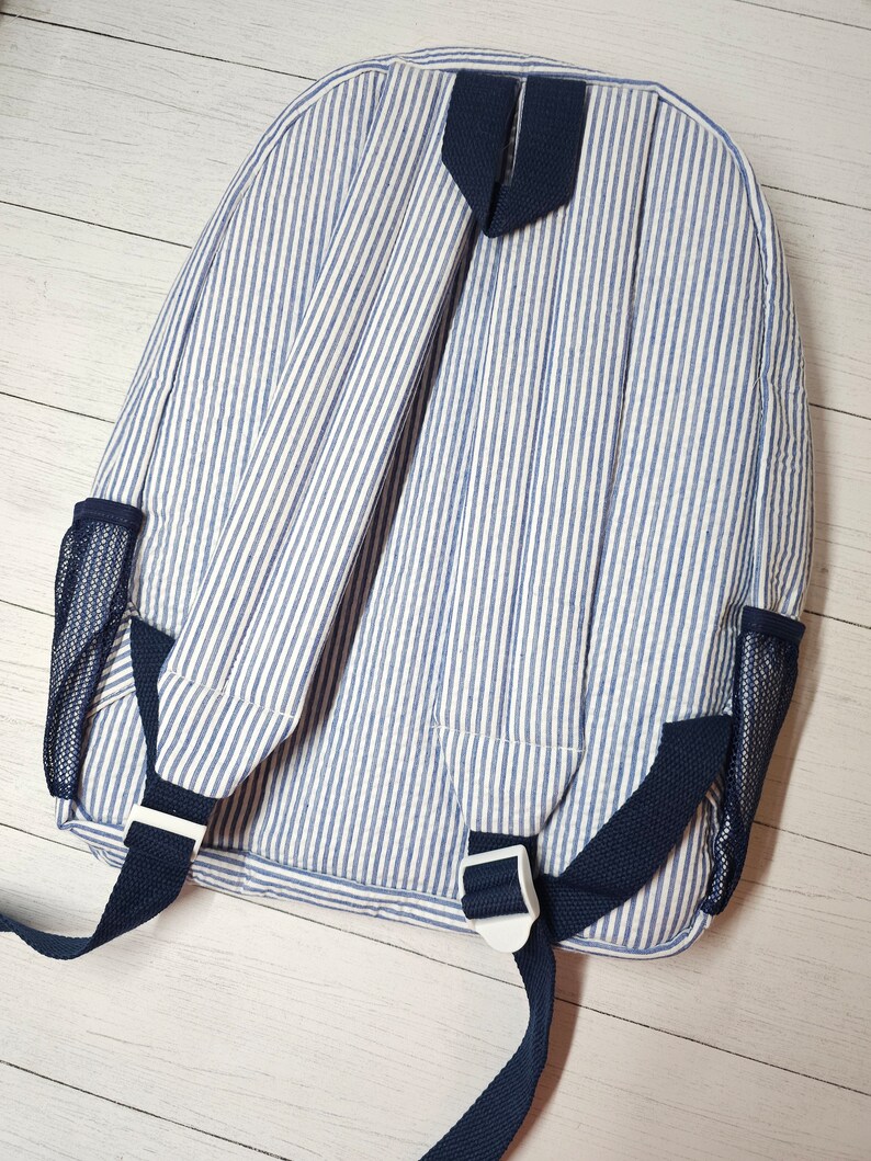 May include: A blue and white striped backpack with navy blue straps and mesh side pockets. The backpack has adjustable straps and white plastic buckles. The fabric has a vertical stripe pattern.