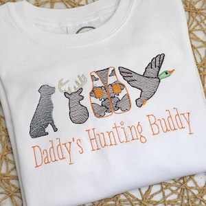 Puede incluir: Mono blanco para bebé con bordado naranja de un chaleco de caza, un ciervo, un perro y un pato. El texto "Daddy's Hunting Buddy" está bordado en naranja debajo de las imágenes.