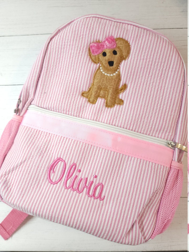 May include: Pink and white striped backpack with a brown dog applique wearing a pink bow and a pearl necklace. The backpack has a pink zipper and the name "Olivia" is embroidered on the front.