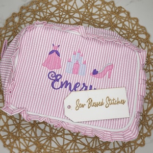 May include: Pink and white striped cosmetic bag with embroidered princess-themed designs, including a dress, castle, and high heel. The name "Emeri" is embroidered in purple script. A tag reads "Sew Blessed Stitches."