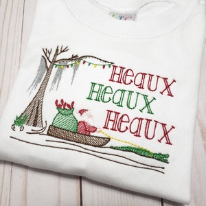 May include: A white t-shirt featuring an embroidered design of Santa Claus in a pirogue boat pulled by an alligator. The scene includes a cypress tree with Spanish moss and string lights. The text "HEAUX HEAUX HEAUX" is stitched in red and green.