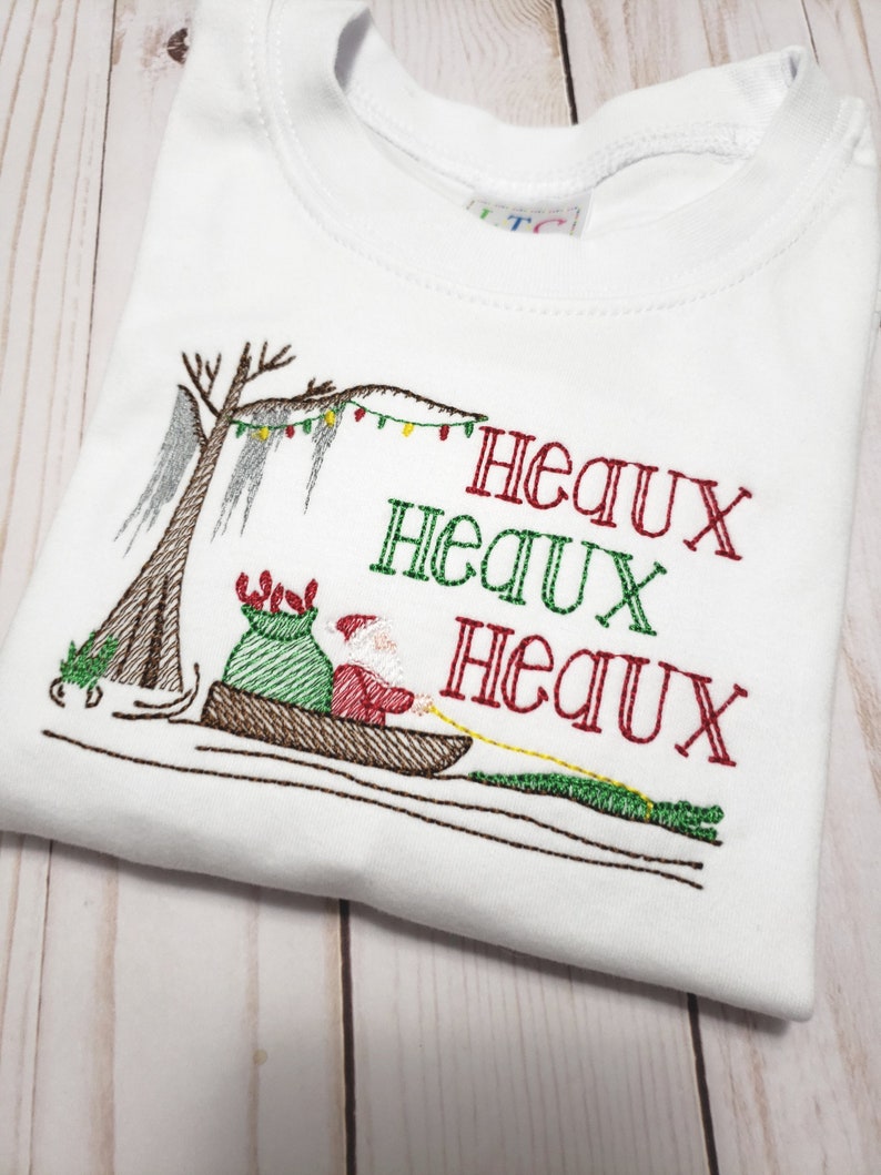 May include: A white t-shirt featuring an embroidered design of Santa Claus in a pirogue boat pulled by an alligator. The scene includes a cypress tree with Spanish moss and string lights. The text "HEAUX HEAUX HEAUX" is stitched in red and green.