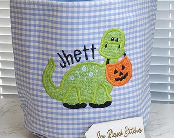 May include: A blue and white gingham fabric basket with a green dinosaur design holding an orange jack-o'-lantern. The name "Jhett" is embroidered above the dinosaur. The basket has a blue interior and a white tag that says "Sew Blessed Stitches".
