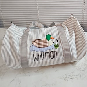 May include: A striped duffel bag with a duck applique. The duck has a green head, brown body, and orange beak. The bag has gray trim and handles, and the name "Whitman" is embroidered below the duck.